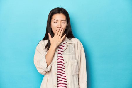 Foto de Mujer asiática con camisa en capas y camiseta a rayas, bostezando mostrando un gesto cansado cubriendo la boca con la mano. - Imagen libre de derechos