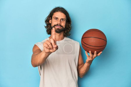 Foto de Hombre barbudo con baloncesto en estudio azul mostrando el número uno con el dedo. - Imagen libre de derechos