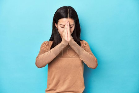 Téléchargez les photos : Jeune femme asiatique au col roulé brun, tenant la main dans la prière près de la bouche, se sent confiant. - en image libre de droit