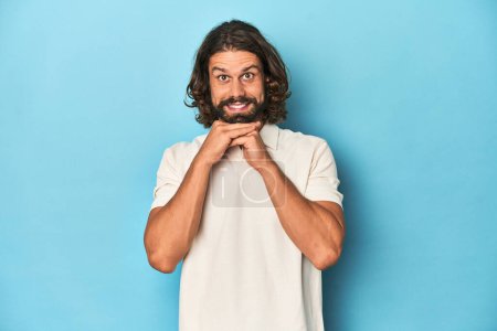Photo for Long-haired man in a white polo, blue studio keeps hands under chin, is looking happily aside. - Royalty Free Image