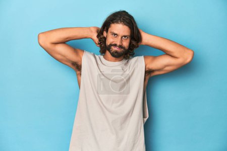 Photo for Bearded man in a tank top, blue backdrop feeling confident, with hands behind the head. - Royalty Free Image