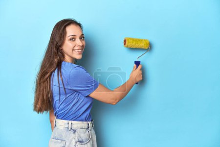 Téléchargez les photos : Jeune femme caucasienne avec rouleau de peinture jaune sur studio bleu. - en image libre de droit