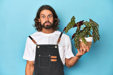Photo for Long-haired gardener with beard holding a plant shrugs shoulders and open eyes confused. - Royalty Free Image
