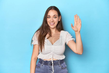 Foto de Mujer joven con estilo en blusa blanca en un fondo de estudio azul sonriente alegre mostrando el número cinco con los dedos. - Imagen libre de derechos
