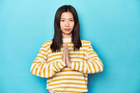 Photo for Asian woman in striped yellow sweater, praying, showing devotion, religious person looking for divine inspiration. - Royalty Free Image