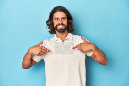Téléchargez les photos : Homme aux cheveux longs dans un polo blanc, studio bleu surpris pointant du doigt, souriant largement. - en image libre de droit