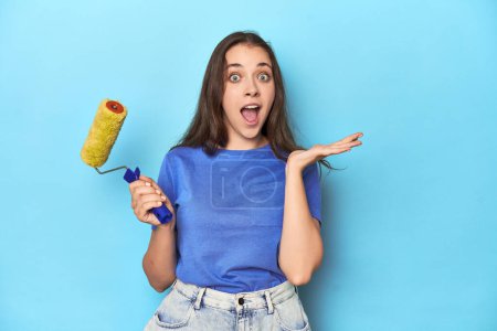 Téléchargez les photos : Jeune femme avec rouleau de peinture jaune sur un fond bleu surpris et choqué. - en image libre de droit
