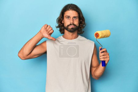 Photo for Bearded man painting with a yellow roller showing a dislike gesture, thumbs down. Disagreement concept. - Royalty Free Image