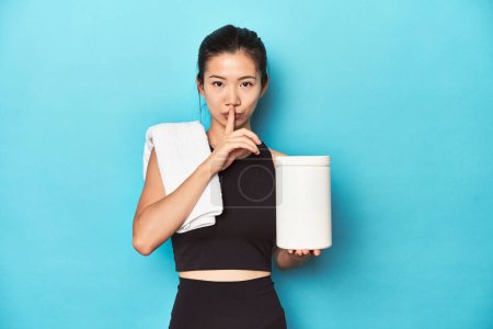 Photo for Young Asian sportswoman with protein bottle, gym setup, keeping a secret or asking for silence. - Royalty Free Image