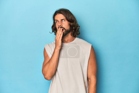 Photo for Bearded man in a tank top, blue backdrop yawning showing a tired gesture covering mouth with hand. - Royalty Free Image