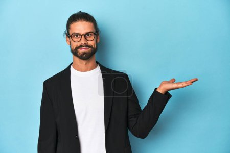 Photo for Businessman in suit with eyeglasses and beard showing a copy space on a palm and holding another hand on waist. - Royalty Free Image