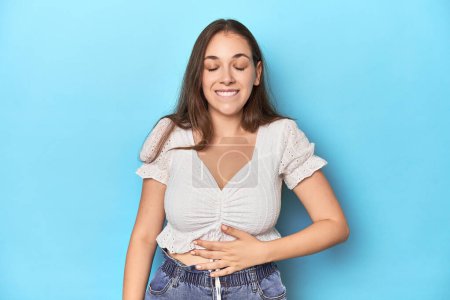 Téléchargez les photos : Jeune femme élégante en chemisier blanc sur fond de studio bleu touche ventre, sourit doucement, concept de manger et de satisfaction. - en image libre de droit