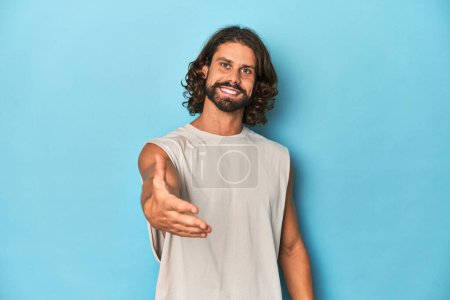 Foto de Hombre barbudo en una camiseta sin mangas, fondo azul estirando la mano a la cámara en gesto de saludo. - Imagen libre de derechos