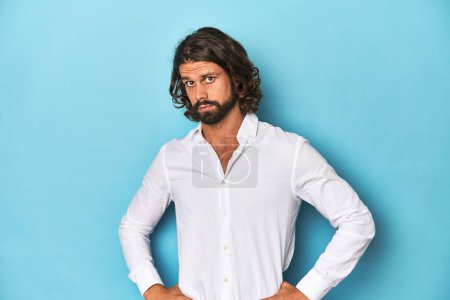 Bearded man in a white shirt, blue backdrop confused, feels doubtful and unsure.