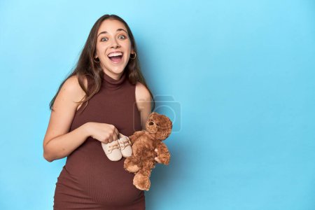 Photo for Pregnant woman holding a baby toy and shoes on blue studio. - Royalty Free Image