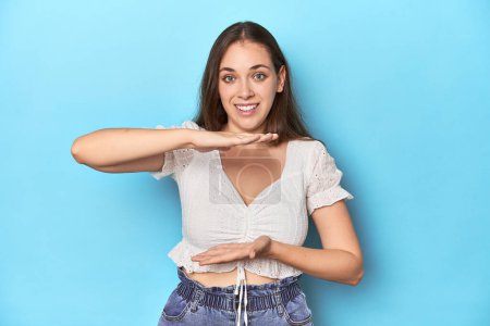 Photo for Stylish young woman in white blouse on a blue studio backdrop holding something with both hands, product presentation. - Royalty Free Image