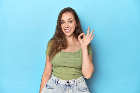 Photo for Young Caucasian woman in a green top on a blue backdrop cheerful and confident showing ok gesture. - Royalty Free Image