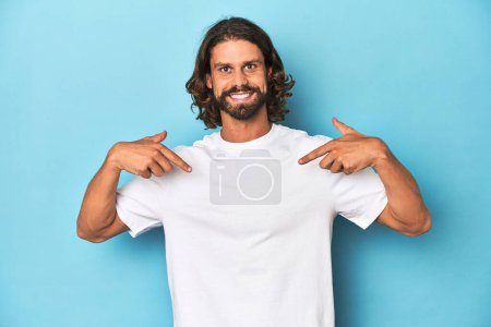Téléchargez les photos : Homme barbu dans une chemise blanche, fond bleu surpris pointant du doigt, souriant largement. - en image libre de droit