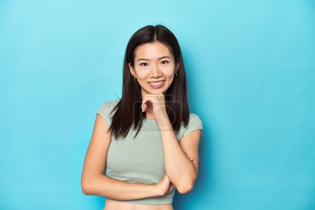 Photo for Asian woman in summer green top, studio backdrop, smiling happy and confident, touching chin with hand. - Royalty Free Image