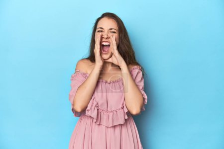 Téléchargez les photos : Jeune femme portant une robe rose sur fond de studio bleu criant excité à l'avant. - en image libre de droit