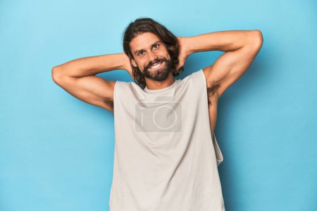 Foto de Hombre barbudo en una camiseta sin mangas, fondo azul brazos extendidos, posición relajada. - Imagen libre de derechos