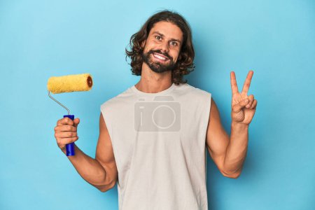 Photo for Bearded man painting with a yellow roller joyful and carefree showing a peace symbol with fingers. - Royalty Free Image