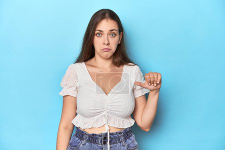 Photo for Stylish young woman in white blouse on a blue studio backdrop showing a dislike gesture, thumbs down. Disagreement concept. - Royalty Free Image