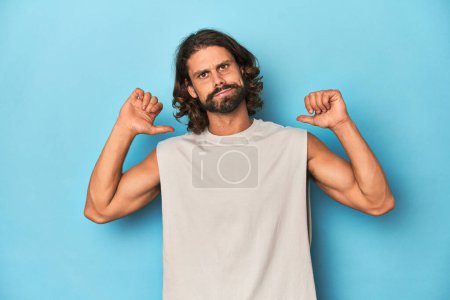 Bearded man in a tank top, blue backdrop feels proud and self confident, example to follow.