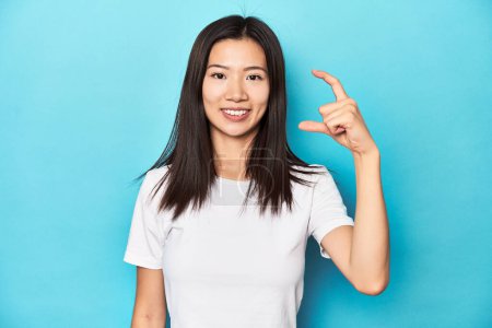 Foto de Mujer asiática joven en camiseta blanca, tiro al estudio, sosteniendo algo poco con los antepasados, sonriendo y confiado. - Imagen libre de derechos