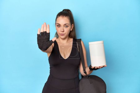 Foto de Mujer joven sosteniendo la botella de proteína en un entorno deportivo de pie con la mano extendida mostrando señal de stop, lo que le impide. - Imagen libre de derechos