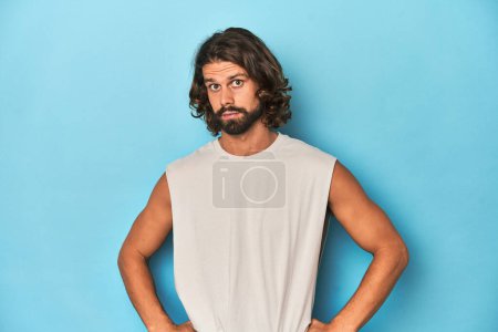Photo for Bearded man in a tank top, blue backdrop confused, feels doubtful and unsure. - Royalty Free Image