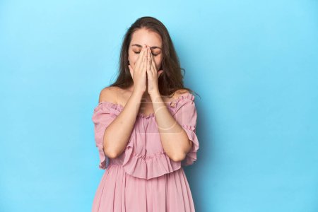 Téléchargez les photos : Jeune femme portant une robe rose sur fond de studio bleu tenant la main dans la prière près de la bouche, se sent confiant. - en image libre de droit