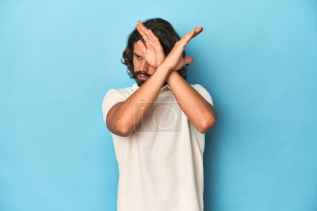 Téléchargez les photos : Homme aux cheveux longs dans un polo blanc, studio bleu gardant deux bras croisés, concept de déni. - en image libre de droit