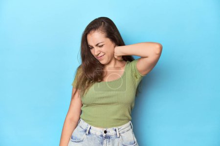 Photo for Young Caucasian woman in a green top on a blue backdrop suffering neck pain due to sedentary lifestyle. - Royalty Free Image