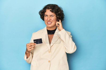 Foto de Mujer joven con tarjeta de crédito, elegante chaqueta blanca que cubre las orejas con las manos. - Imagen libre de derechos
