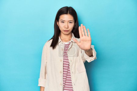 Foto de Mujer asiática con camisa en capas y camiseta a rayas, de pie con la mano extendida mostrando señal de stop, lo que le impide. - Imagen libre de derechos