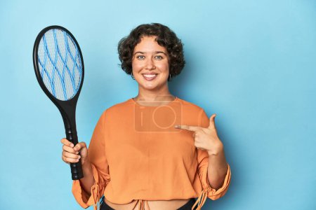 Téléchargez les photos : Jeune femme avec raquette moustique électrique Jeune femme avec raquette moustique électrique pointant à la main vers un espace de copie de chemise, fière et confiante - en image libre de droit