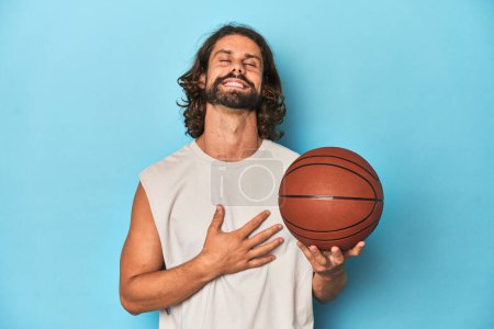Téléchargez les photos : Homme barbu avec basket en studio bleu rit bruyamment en gardant la main sur la poitrine. - en image libre de droit