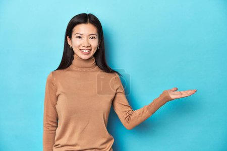 Photo for Young Asian woman in brown turtleneck, showing a copy space on a palm and holding another hand on waist. - Royalty Free Image