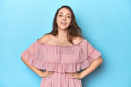 Photo for Young woman wearing a pink dress on a blue studio backdrop confident keeping hands on hips. - Royalty Free Image
