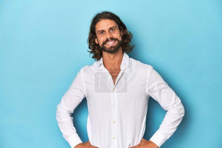 Photo for Bearded man in a white shirt, blue backdrop confident keeping hands on hips. - Royalty Free Image
