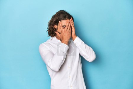 Photo for Bearded man in a white shirt, blue backdrop blink through fingers frightened and nervous. - Royalty Free Image