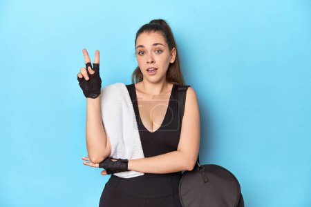 Téléchargez les photos : Jeune femme prête pour la gym avec des éléments essentiels sur fond bleu montrant numéro deux avec les doigts. - en image libre de droit