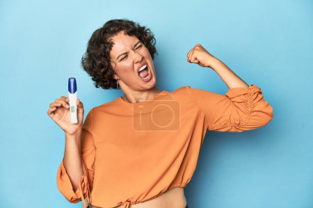Téléchargez les photos : Jeune femme tenant un test de grossesse, arrière-plan studio levant le poing après une victoire, concept gagnant. - en image libre de droit