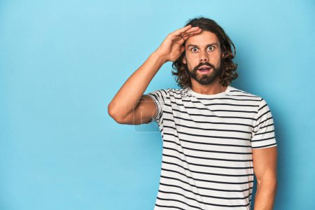 Photo for Bearded man in a striped shirt, blue backdrop shouts loud, keeps eyes opened and hands tense. - Royalty Free Image