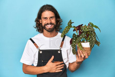 Foto de Jardinero de pelo largo con barba sosteniendo una planta se ríe en voz alta manteniendo la mano en el pecho. - Imagen libre de derechos