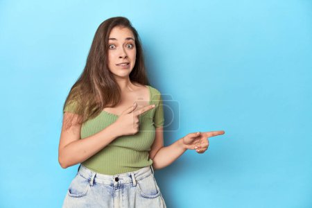 Foto de Joven mujer caucásica en una parte superior verde sobre un fondo azul impactado señalando con los dedos índice a un espacio de copia. - Imagen libre de derechos