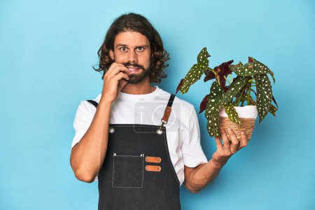 Foto de Jardinero de pelo largo con barba sosteniendo una planta mordiendo las uñas, nervioso y muy ansioso. - Imagen libre de derechos