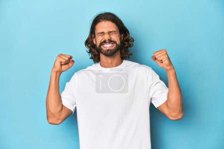 Foto de Hombre barbudo con una camisa blanca, fondo azul animando despreocupado y emocionado. Concepto de victoria. - Imagen libre de derechos