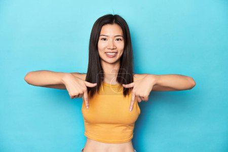 Photo for Asian woman in summer yellow top, studio setup, points down with fingers, positive feeling. - Royalty Free Image